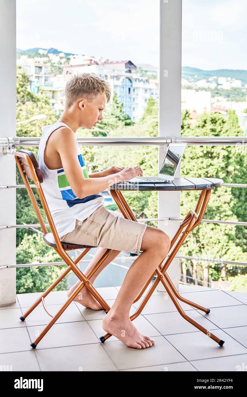Un ragazzo che si siede sul balcone dell'hotel e naviga in internet su un computer portatile grigio. Lo scolaro ama trascorrere le vacanze estive e giocare Foto Stock