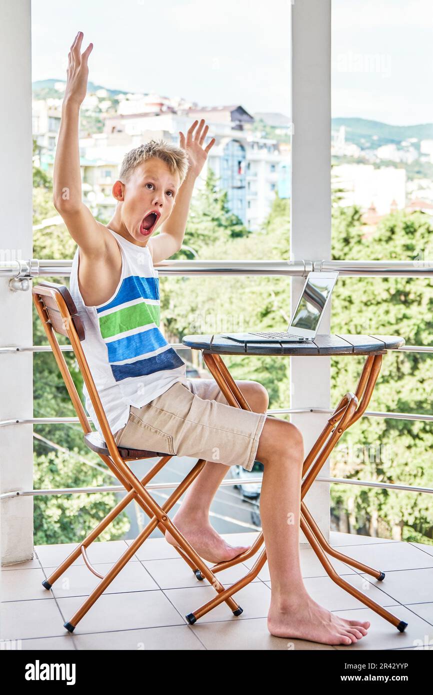 Il ragazzo che gioca un interessante videogioco sul laptop grigio celebra la vittoria. Lo scolaro trascorre le vacanze estive seduti al tavolo sul balcone dell'hotel Foto Stock