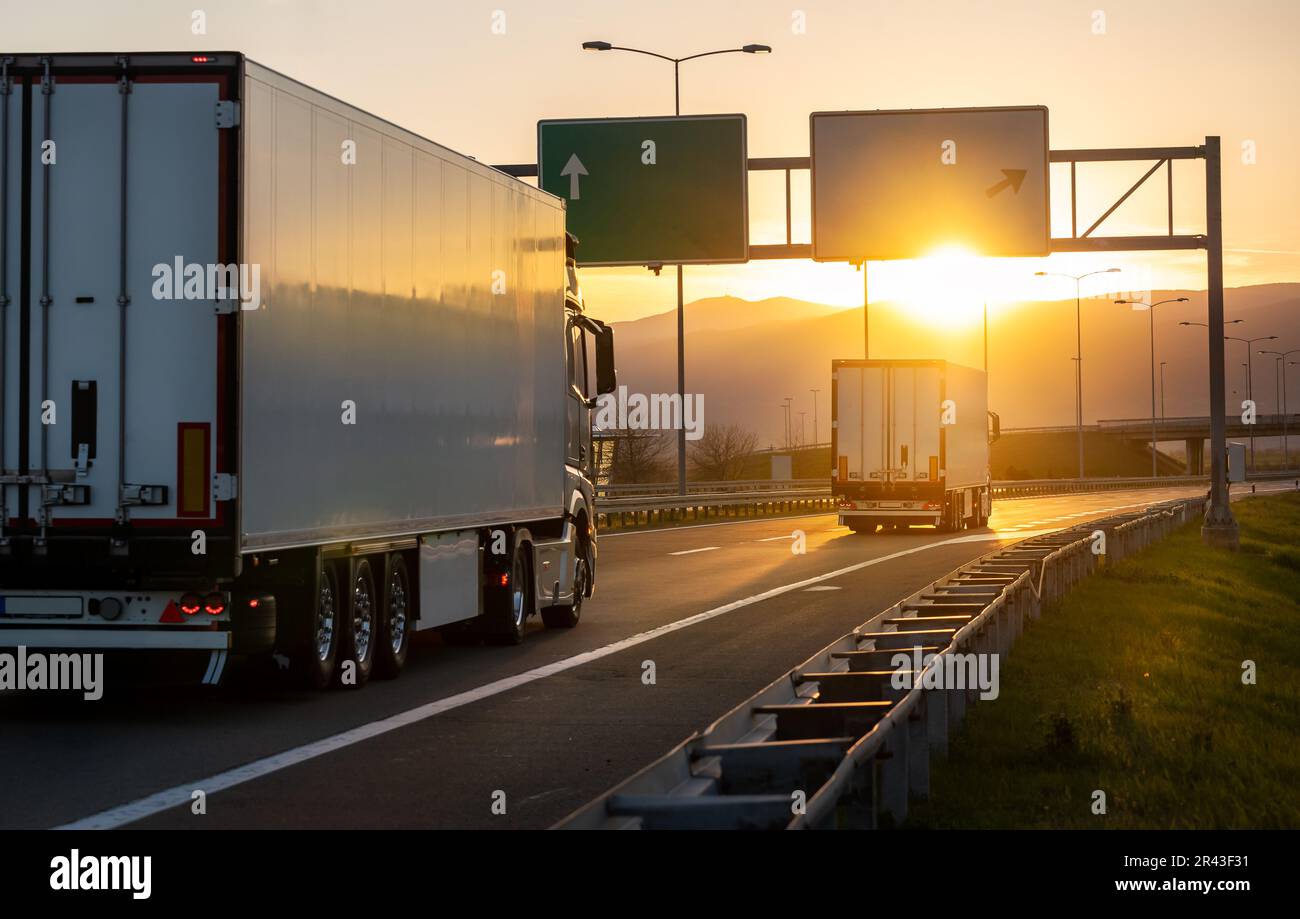 Vista dall'angolo posteriore dei camion di carico per la consegna sulla strada che va verso ovest con il tramonto sullo sfondo. Consegna rapida, logistica di carico e nave di trasporto Foto Stock
