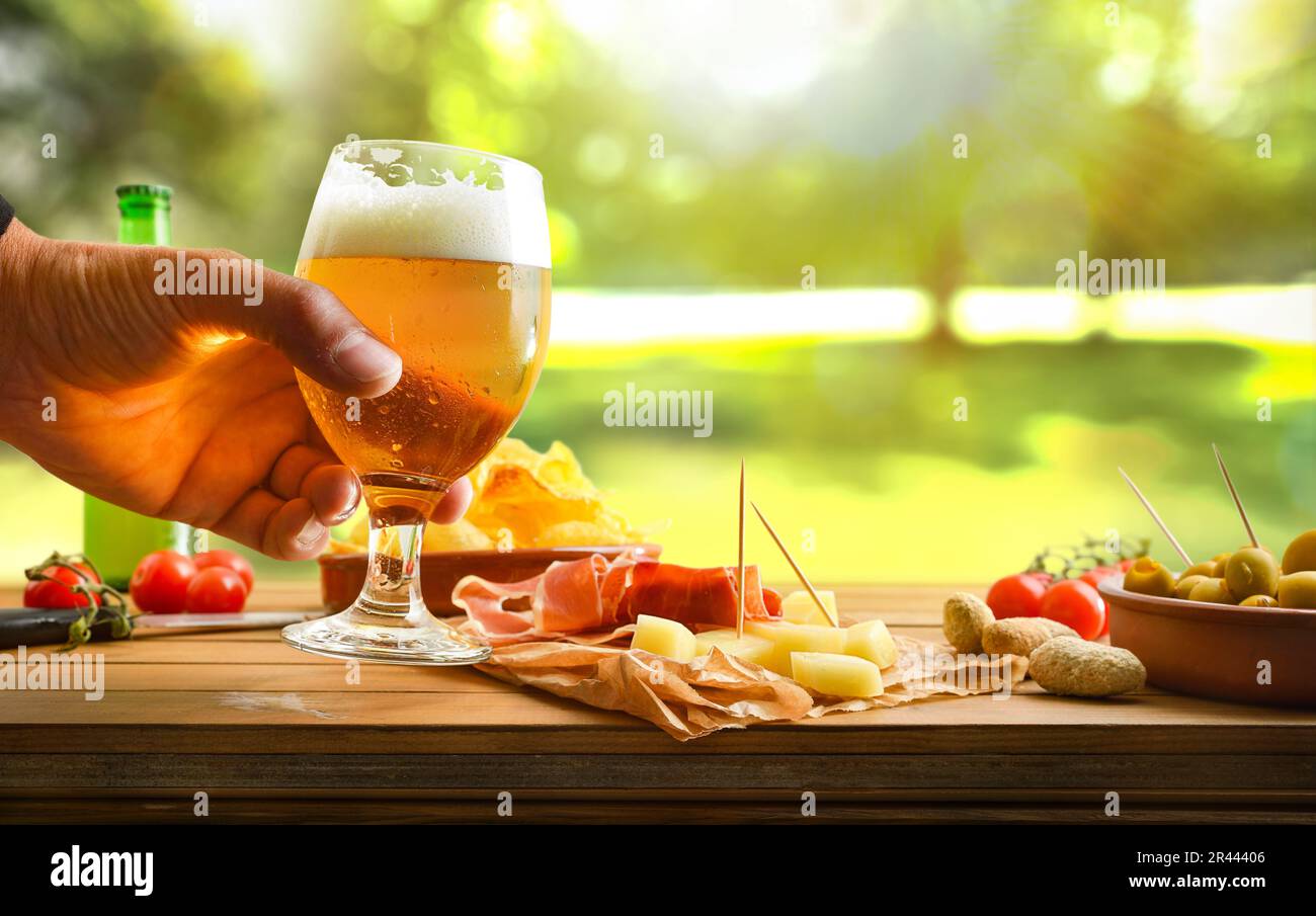 Aperitivo in campagna a mano con un bicchiere di birra su un tavolo di legno con spuntini assortiti e sfondo naturale. Vista frontale. Foto Stock