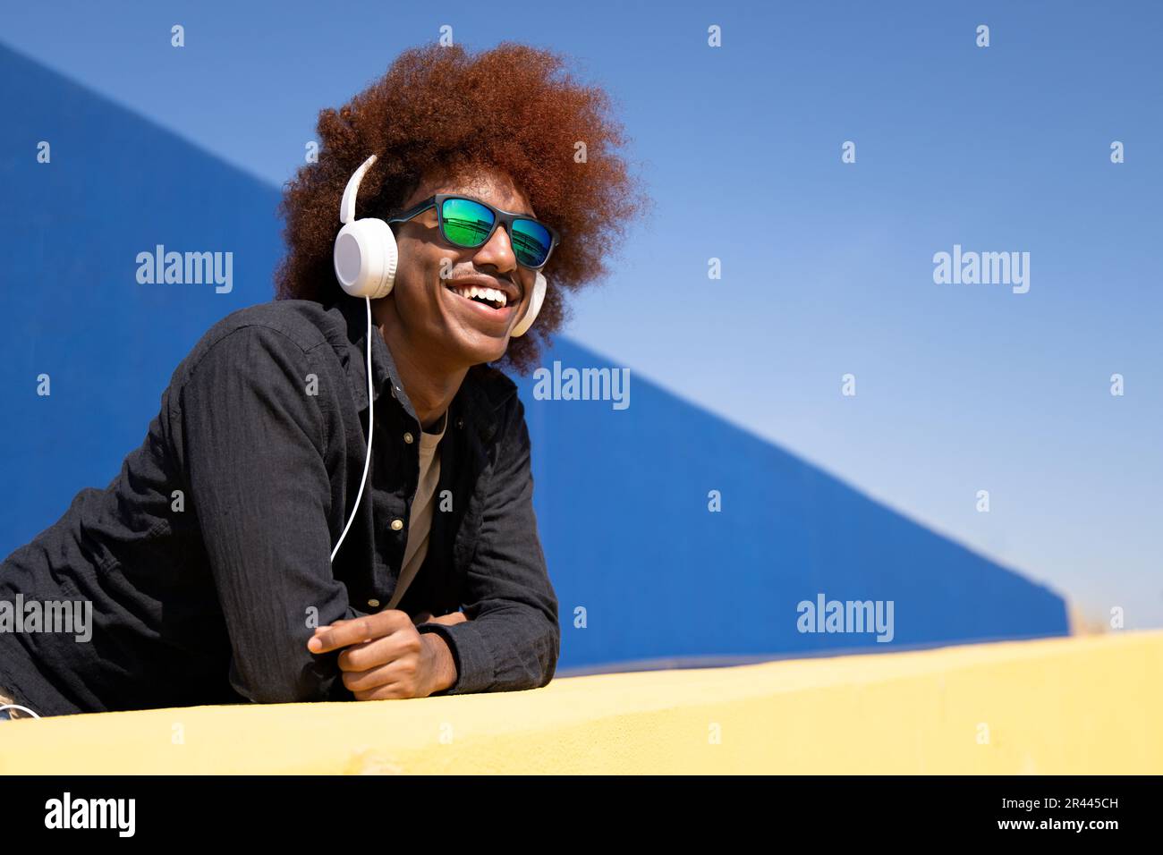 Vista laterale di un giovane uomo gioioso in piedi su una parete che indossa occhiali da sole Foto Stock