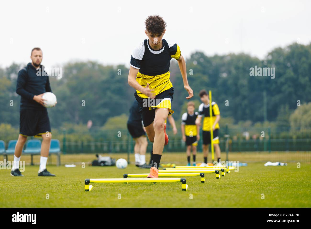 Giovane ragazzo sportivo che salta sugli Hurdles. Unità di pratica di Calcio all'aperto per Club di Calcio Giovanile. Giocatori su allenamento Drill al campo di calcio estivo Foto Stock