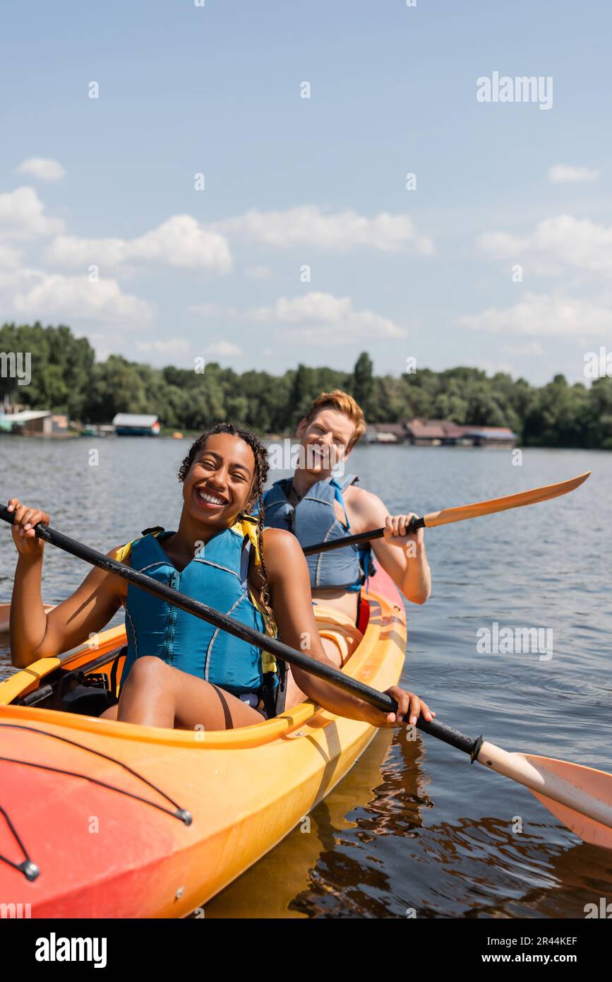 coppia multietnica eccitata in giubbotti di vita trascorrere il fine settimana estivo sul lago pittoresco, mentre pagaia in kayak sportivo sotto il cielo blu con nuvole bianche Foto Stock