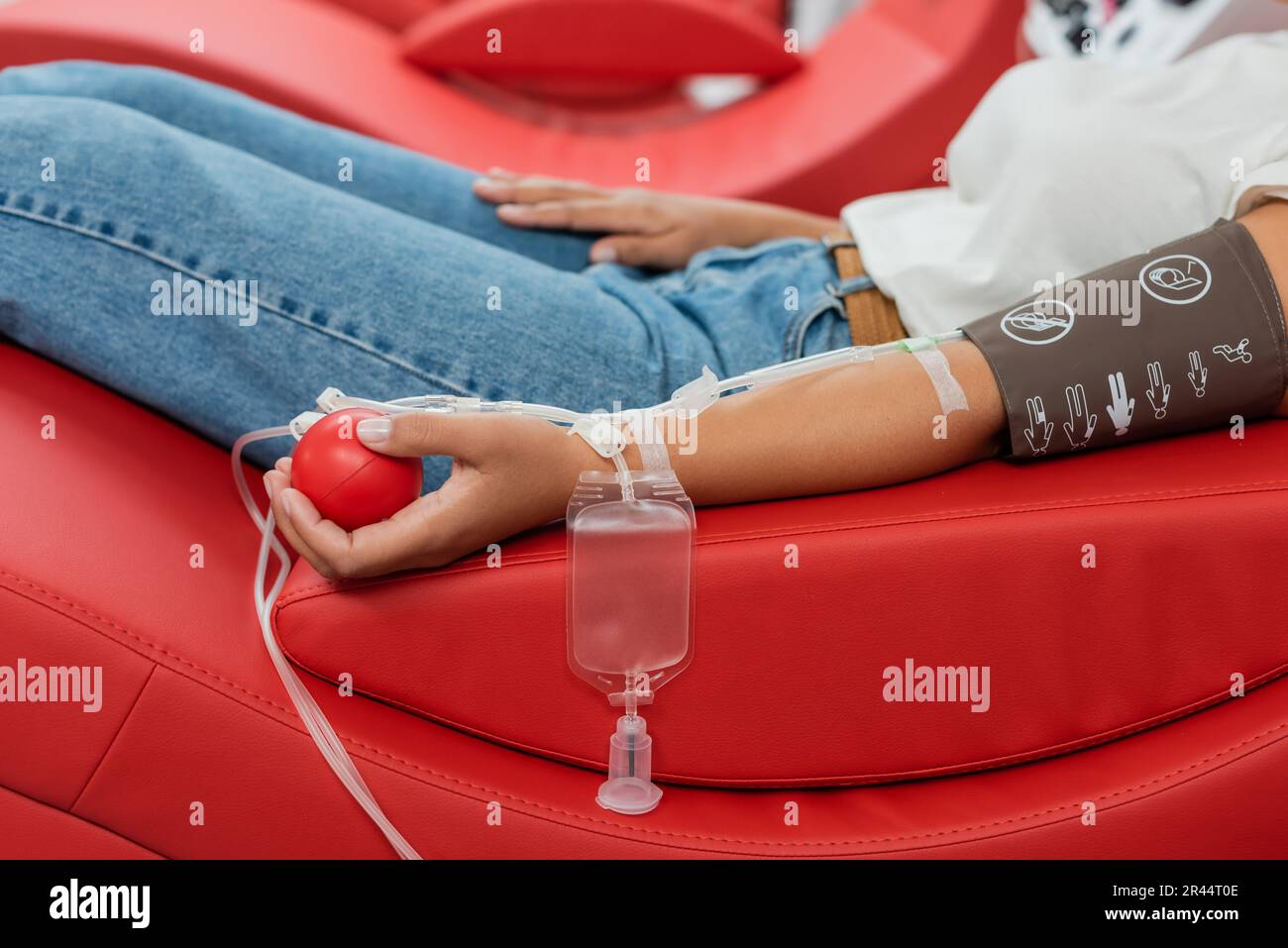 vista parziale della donna multirazziale con set trasfusionale e palla medica di gomma seduta su sedia medica ergonomica durante la donazione di sangue in laboratorio Foto Stock