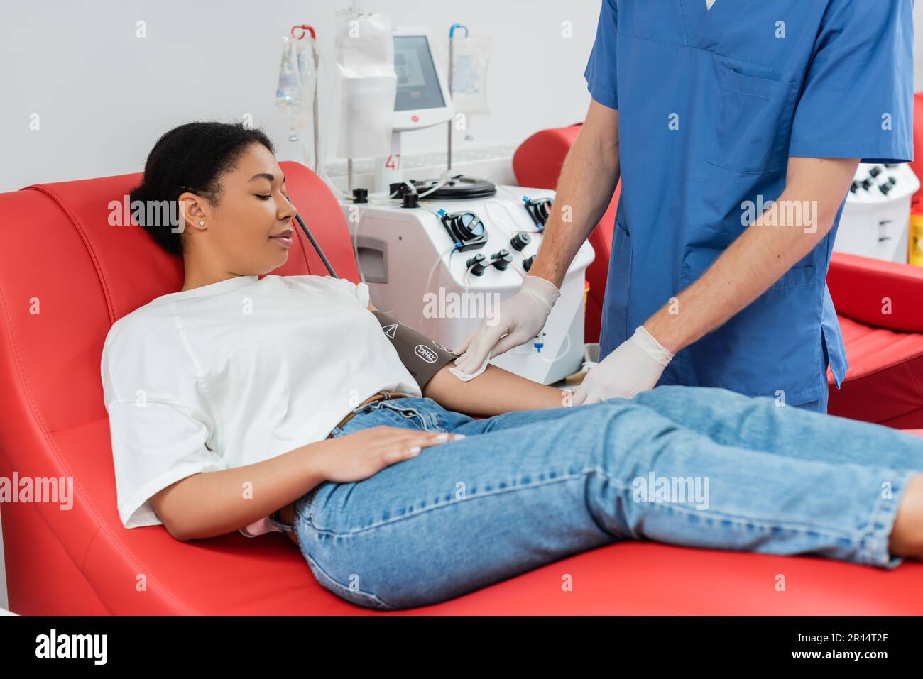 operatore sanitario in uniforme blu e guanti in lattice che tiene tampone alcool e braccio di pulizia multirazziale donna in bracciale pressione del sangue seduta su medico c Foto Stock