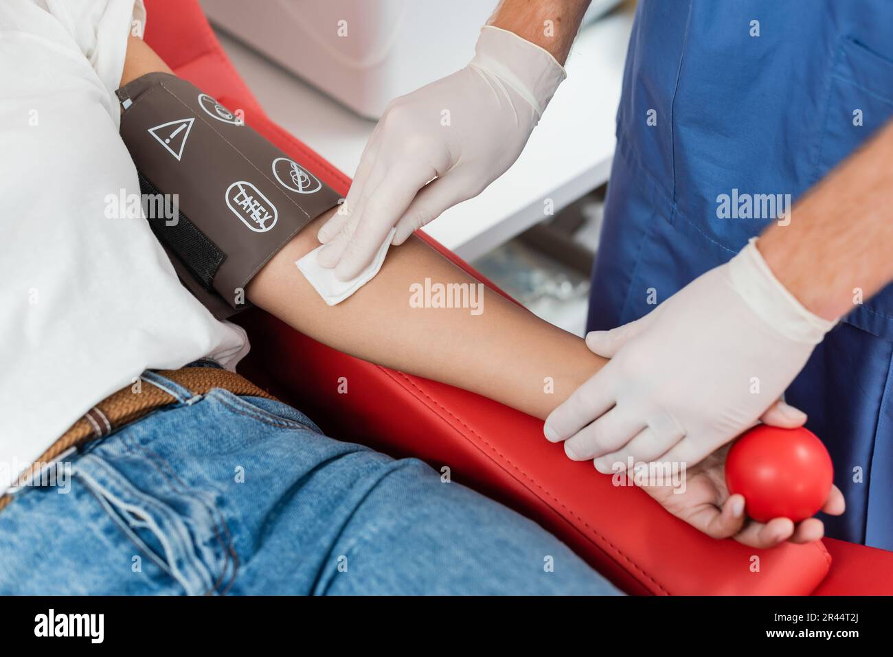 vista ritagliata del medico in guanti di lattice che tengono tampone di alcool e braccio di pulizia di donna multirazziale che tiene la palla di gomma mentre si siede sulla sedia medica in b Foto Stock
