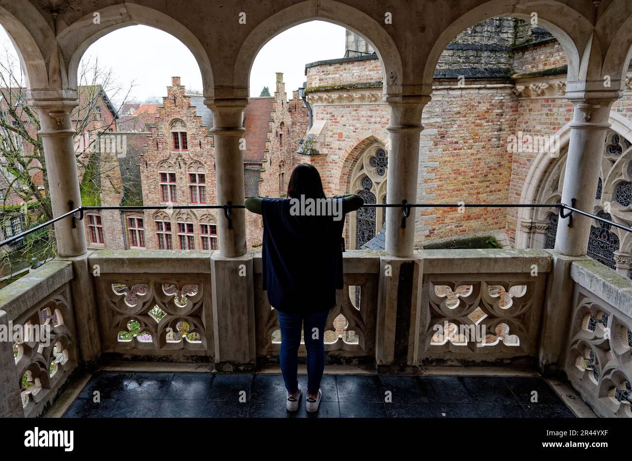 Retro di una giovane donna che indossa un mantello blu che guarda ad un paesaggio urbano medievale da un balcone sopraelevato. Balcone nella città di Brugges. Foto Stock