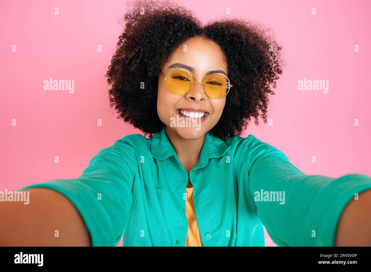 Bella donna alla moda brasiliano o ispanica, blogger, indossare camicia verde casual e occhiali alla moda, scattare foto selfie, guarda la macchina fotografica, sorrisi, stare in piedi su sfondo isolato rosa pastello Foto Stock