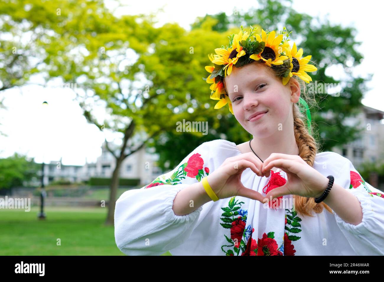 Una donna Ucraina in camicia ricamata con una corona di girasole sulla sua testa fa un cuore con le sue mani riconoscimento approvazione gioia Nadezhda piacevoli emozioni rosso-capelli bellezza sulla strada Foto Stock