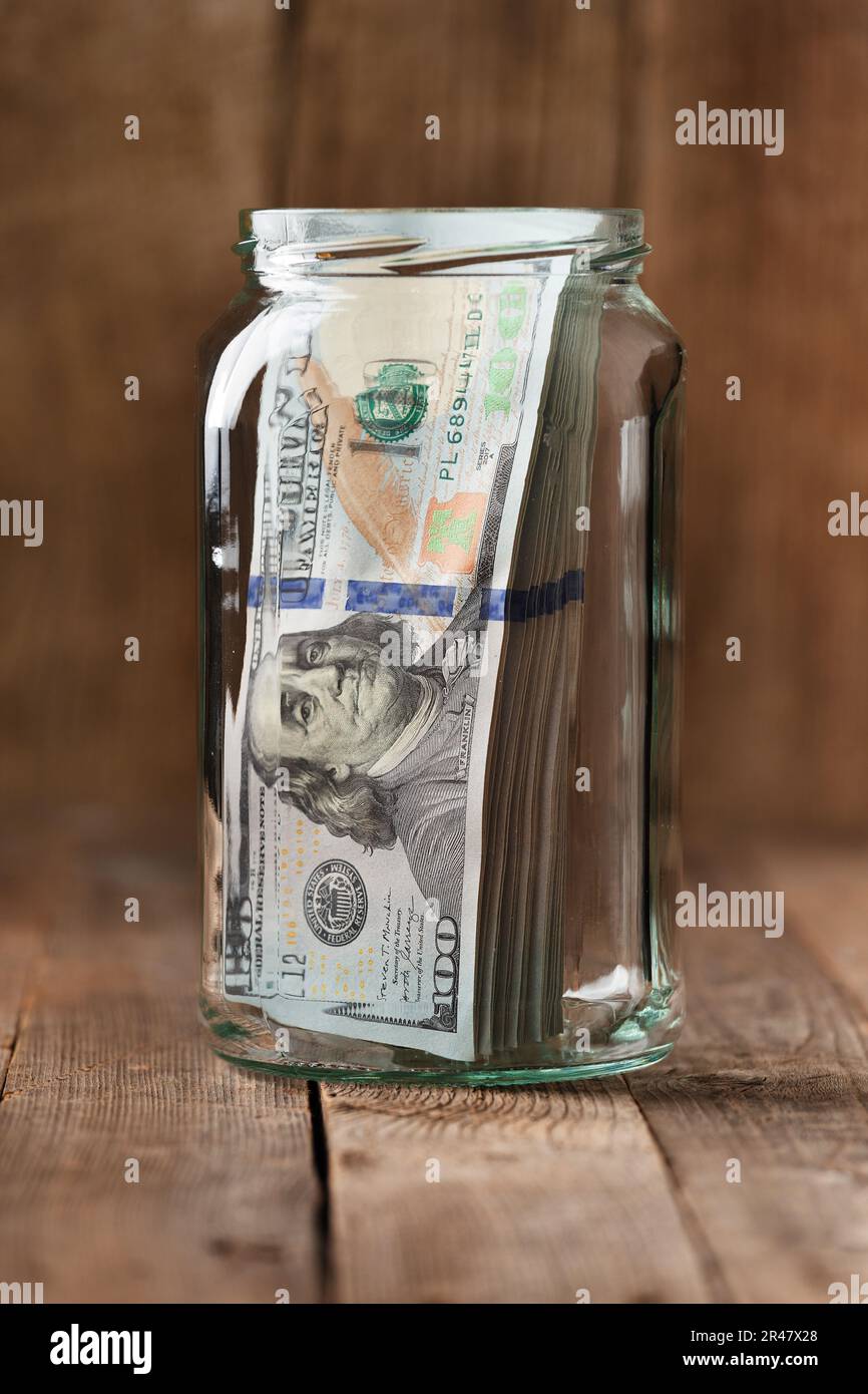 Banconote in dollari in un vaso di vetro su uno sfondo di legno, una pila di banconote da 100 dollari in un vaso. Il concetto di risparmio di denaro, l'accumulo di fondi. Foto Stock
