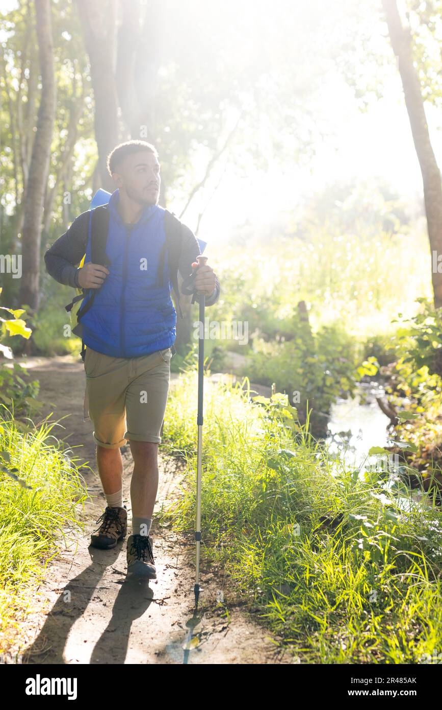 Felice uomo biraciale che indossa zaino e tenendo il palo da trekking, trekking nella foresta, inalterato Foto Stock