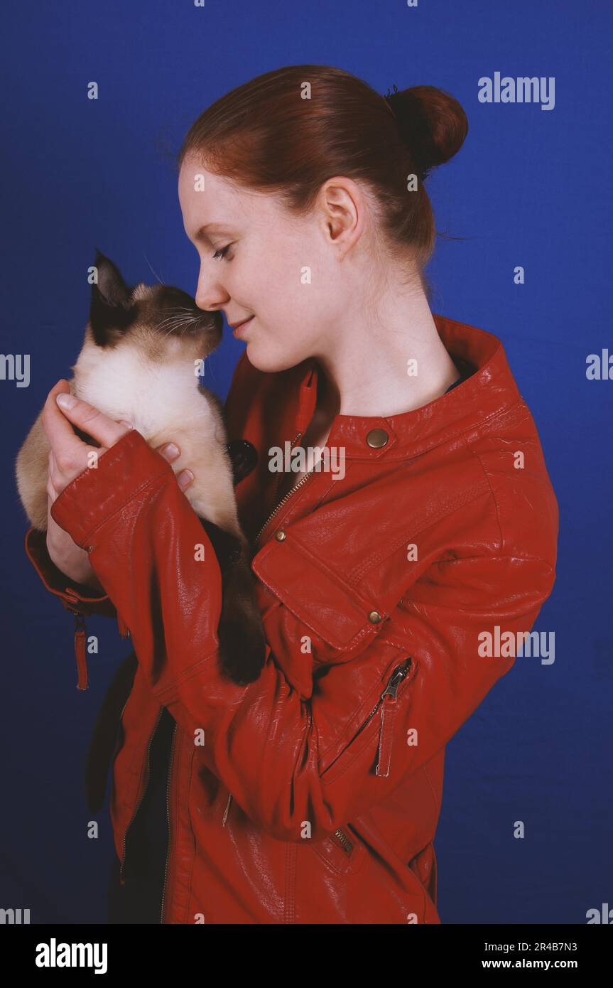giovane donna che indossa una giacca di pelle rossa che tocca il naso con il suo gatto siamese Foto Stock