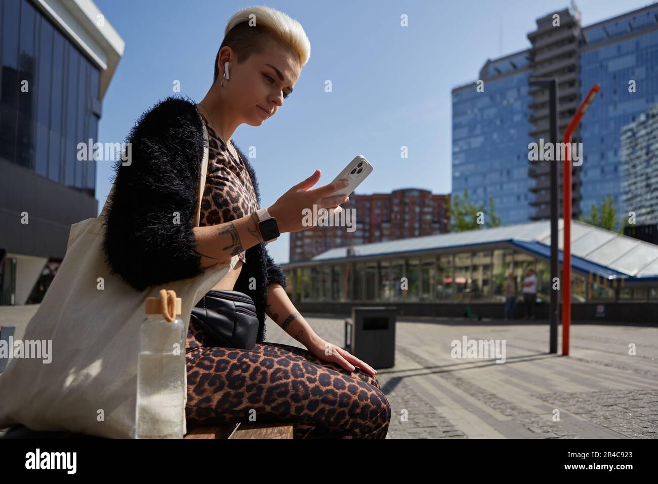 Donna elegante e variegata con capelli corti sbiancati seduti su una panca nel centro della città. Bella donna bianca che indossa cuffie wireless e marrone Foto Stock