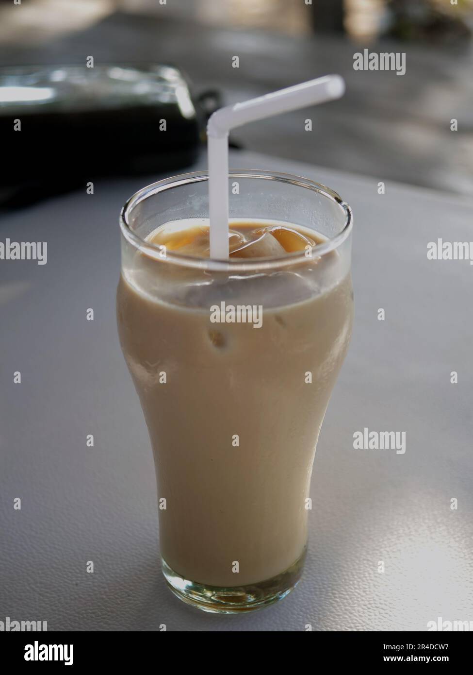 Uno sfondo sfocato e un caffè ghiacciato in un bicchiere su un tavolo di plastica con una cannuccia bianca. Foto Stock
