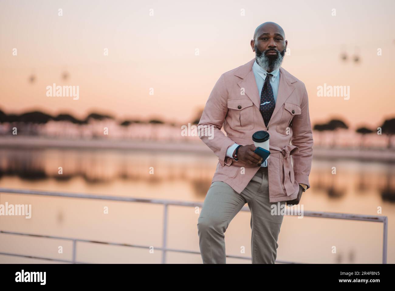 Cielo rosa, sfondo del fiume Tago. Attenzione selettiva a un elegante uomo nero bald maturo in giacca rosa, camicia bianca, pantaloni crema e cravatta di buon gusto. Foto Stock
