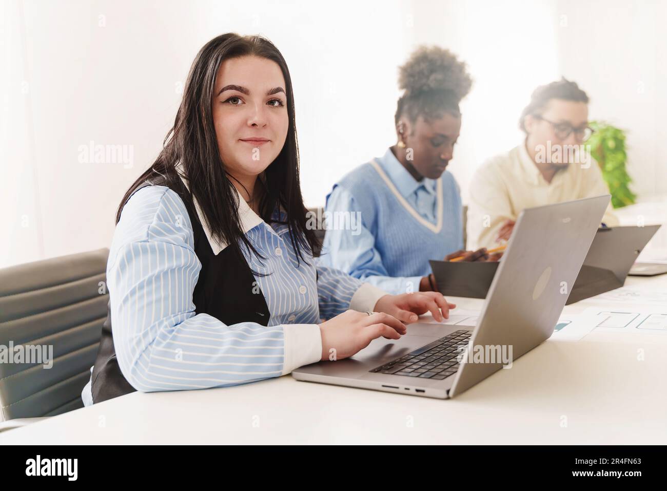 Donna caucasica con piercing al lavoro sul portatile - in un ufficio diverso, una giovane donna si concentra sul suo portatile, i suoi colleghi africani e cinesi sfocati Foto Stock