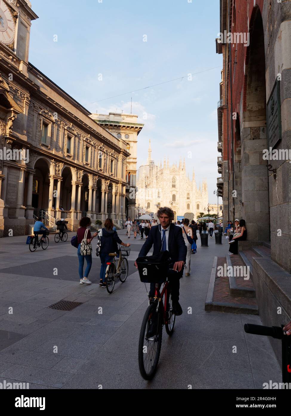Uomo che indossa un abito in bicicletta con un cesto sul davanti in una strada di Milano con il Duomo illuminato dalla luce della sera dietro, Lombardia, Italia Foto Stock