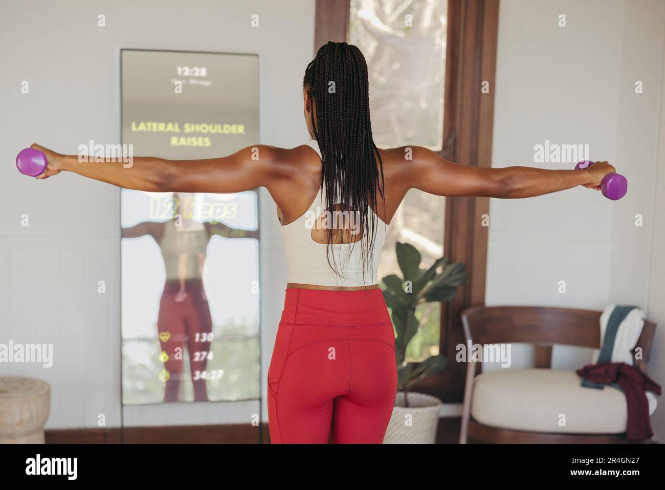 Partecipando a una lezione di allenamento virtuale attraverso uno specchio per il fitness intelligente, una donna aggiunge esercizi a manubrio nella sua routine di allenamento, utilizzando una forma avanzata Foto Stock