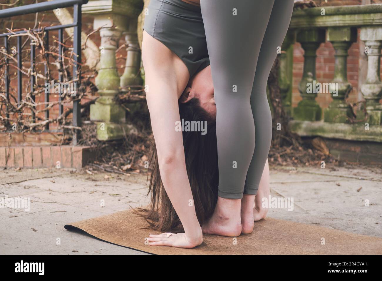 Primo piano di una donna caucasica nuova insegnante di yoga facendo un'uttanasana curva in piedi in avanti indossando abbigliamento sportivo verde sulla terrazza di un paesaggio storico Foto Stock