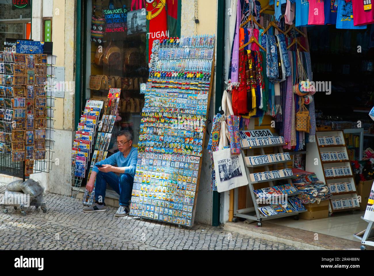 Un uomo che guarda il suo cellulare mentre si siede fuori da un negozio di souvenir turistico a Lisbona Foto Stock