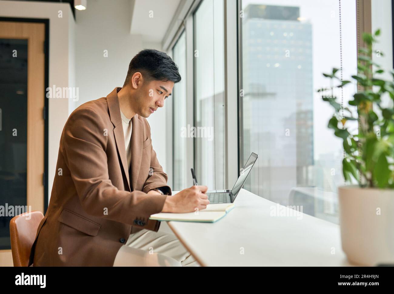 Giovane uomo d'affari asiatico impegnato che lavora su un computer portatile in ufficio scrivendo appunti. Foto Stock