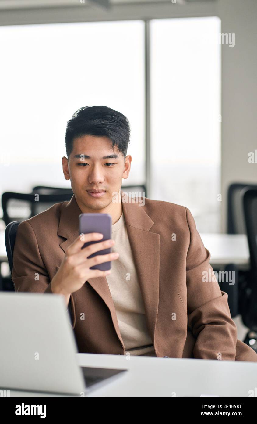 Giovane uomo d'affari asiatico impegnato che utilizza lo smartphone in ufficio. Verticale Foto Stock