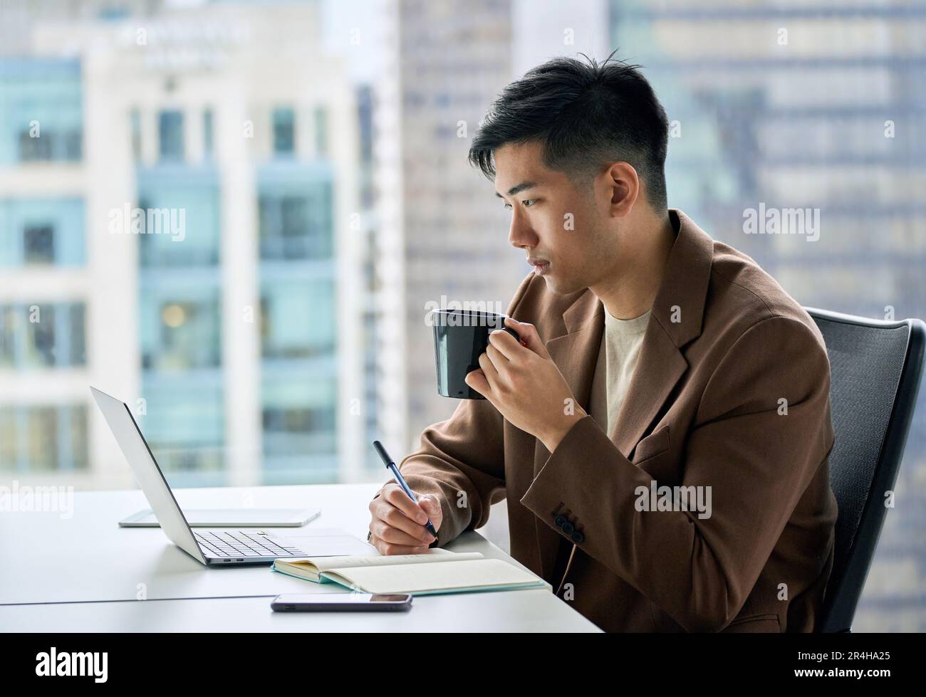 Giovane uomo d'affari asiatico che lavora su un computer portatile in ufficio bevendo caffè. Foto Stock