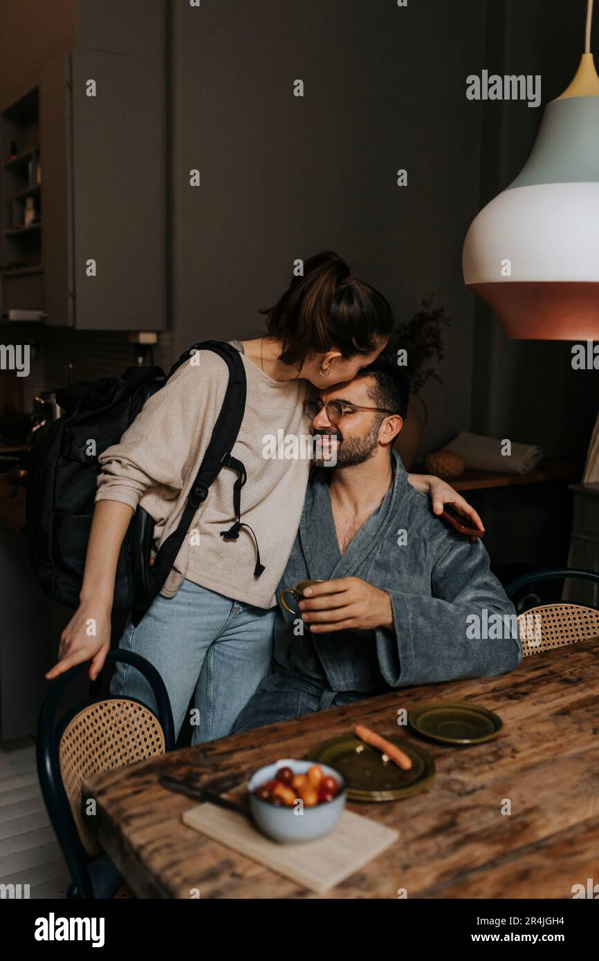 Persona non binaria che bacia il ragazzo seduto al tavolo da pranzo in camera Foto Stock