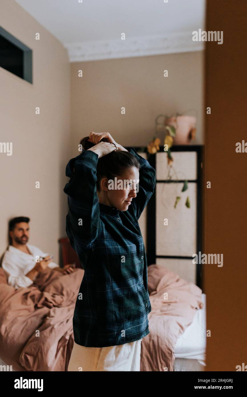 Non-binary persona che legano i capelli mentre si trova in piedi in camera da letto a casa Foto Stock