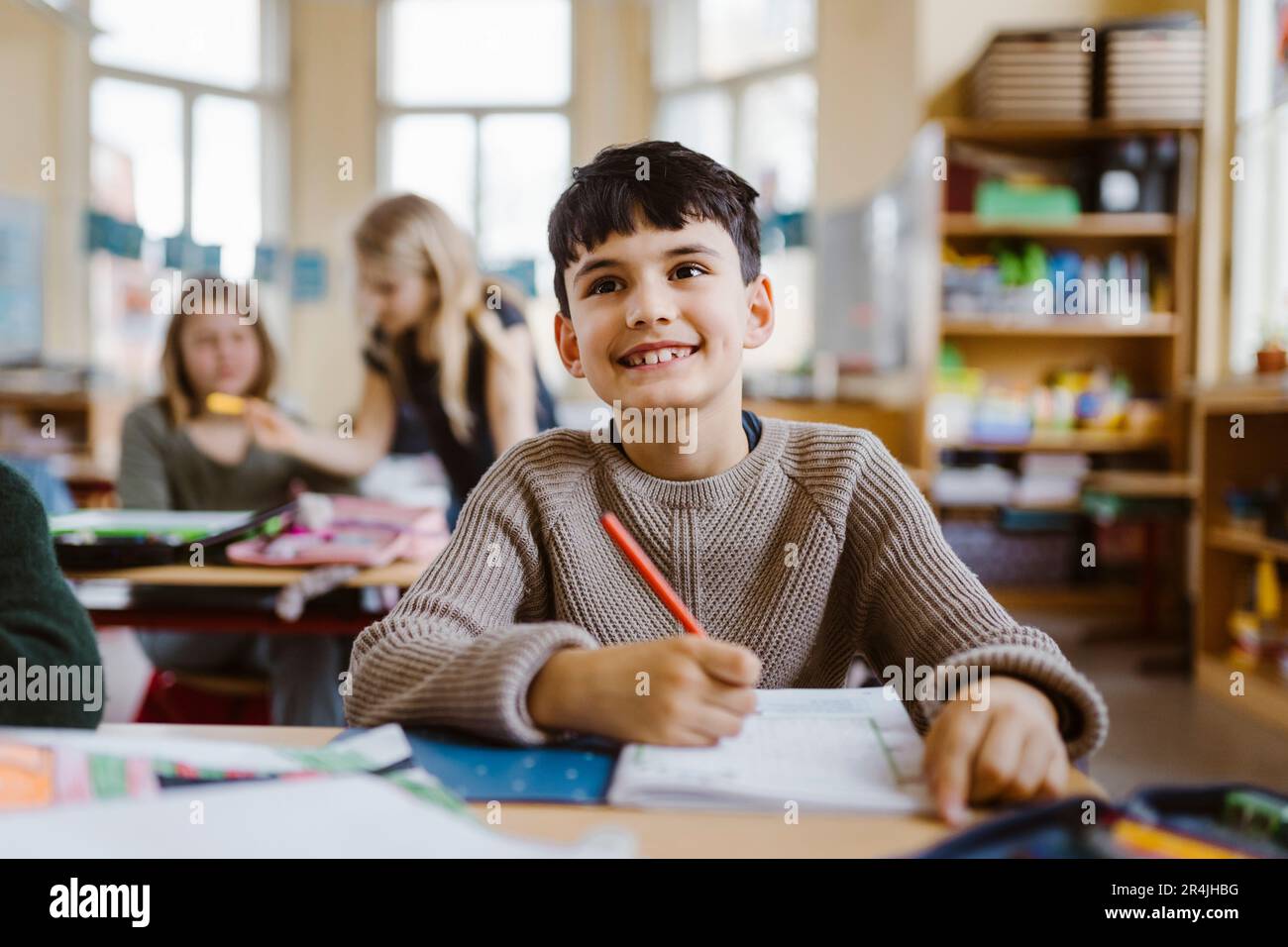 Allievo sorridente che tiene la matita mentre siede alla scrivania in classe Foto Stock