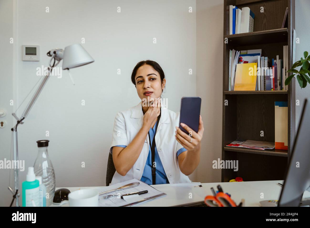 Medico di sesso femminile che effettua una videochiamata online con il paziente tramite smartphone mentre si siede alla scrivania in clinica Foto Stock