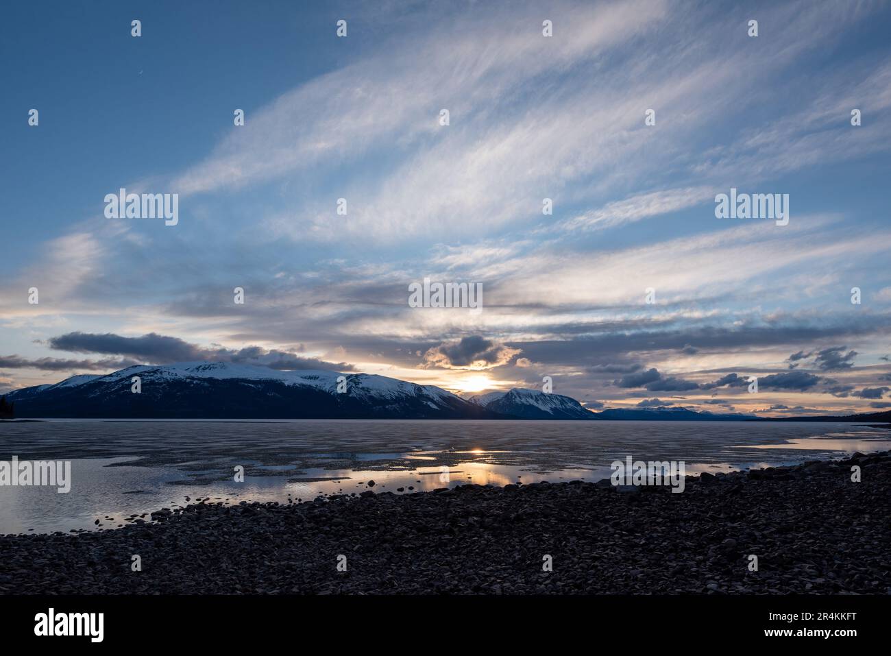 Tramonto roccioso sulla riva del lago ad Atlin, British Columbia durante la primavera. Montagne innevate che si riflettono nella calma acqua del lago sottostante con un cielo spettacolare. Foto Stock