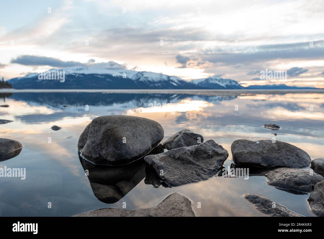 Tramonto roccioso sulla riva del lago ad Atlin, British Columbia durante la primavera. Montagne innevate che si riflettono nella calma acqua del lago sottostante con un cielo spettacolare. Foto Stock
