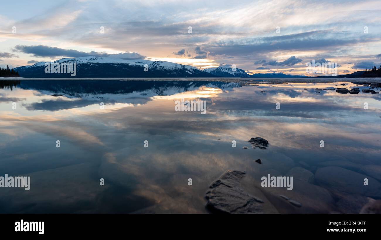 Tramonto roccioso sulla riva del lago ad Atlin, British Columbia durante la primavera. Montagne innevate che si riflettono nella calma acqua del lago sottostante con un cielo spettacolare. Foto Stock