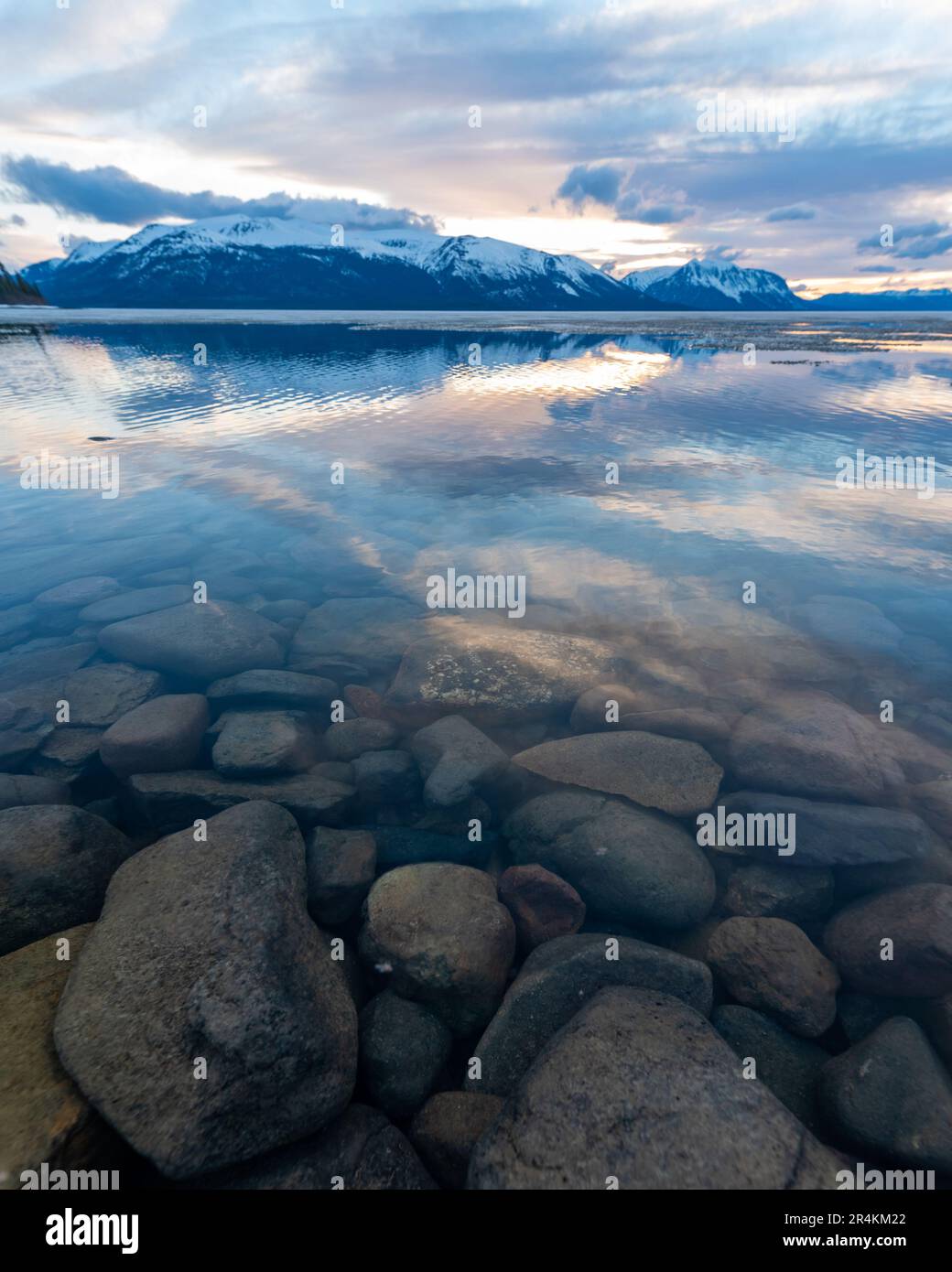 Tramonto roccioso sulla riva del lago ad Atlin, British Columbia durante la primavera. Montagne innevate che si riflettono nella calma acqua del lago sottostante con un cielo spettacolare. Foto Stock