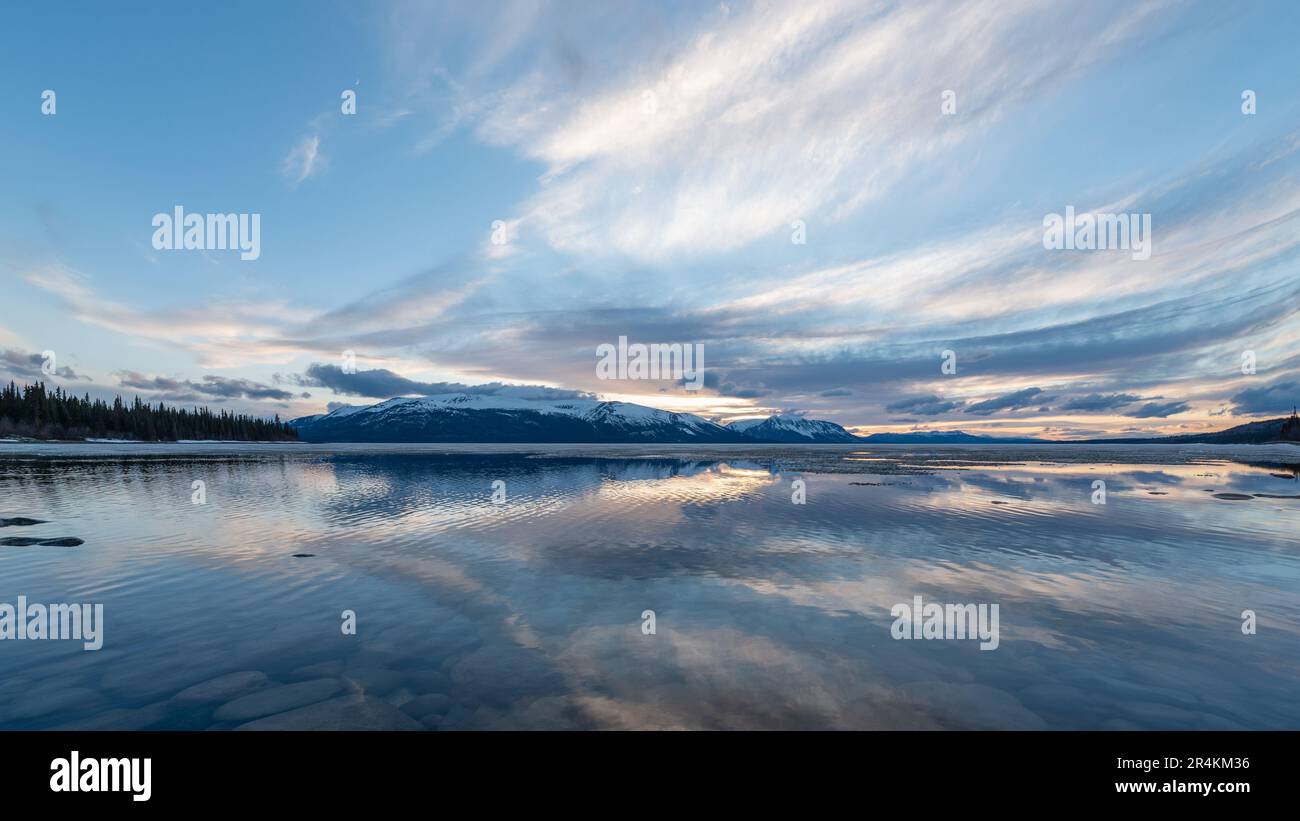 Tramonto roccioso sulla riva del lago ad Atlin, British Columbia durante la primavera. Montagne innevate che si riflettono nella calma acqua del lago sottostante con un cielo spettacolare. Foto Stock