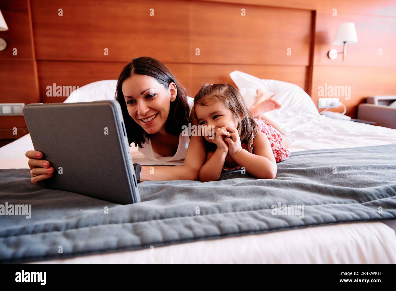 Una bambina e la sua giovane madre si divertono a usare un iPad sul letto della loro camera d'albergo Foto Stock