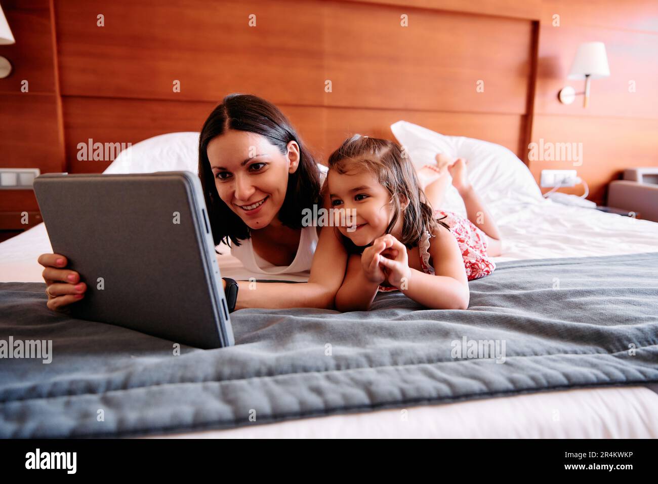 Una bambina e la sua giovane madre si divertono a usare un iPad sul letto della loro camera d'albergo Foto Stock