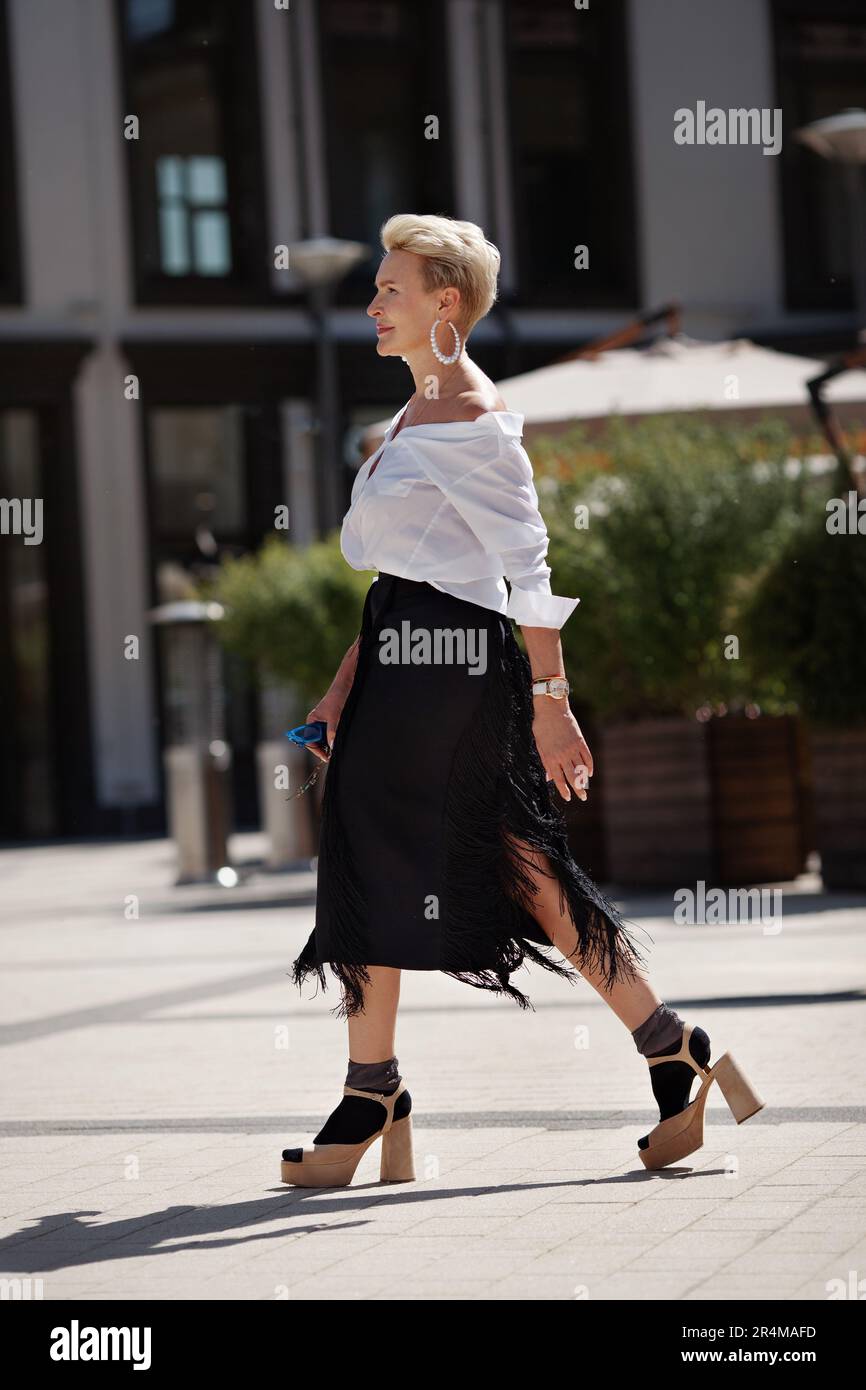 Fiducia e raffinatezza donna matura cammina per le strade della città in  elegante ensemble camicia bianca chic e gonna con frange nere con sandalo  con tacco alto Foto stock - Alamy