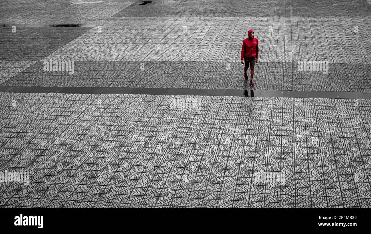 Un uomo in un cappotto rosso cammina attraverso una piazza lastricata sotto la pioggia a Danang, Vietnam Foto Stock