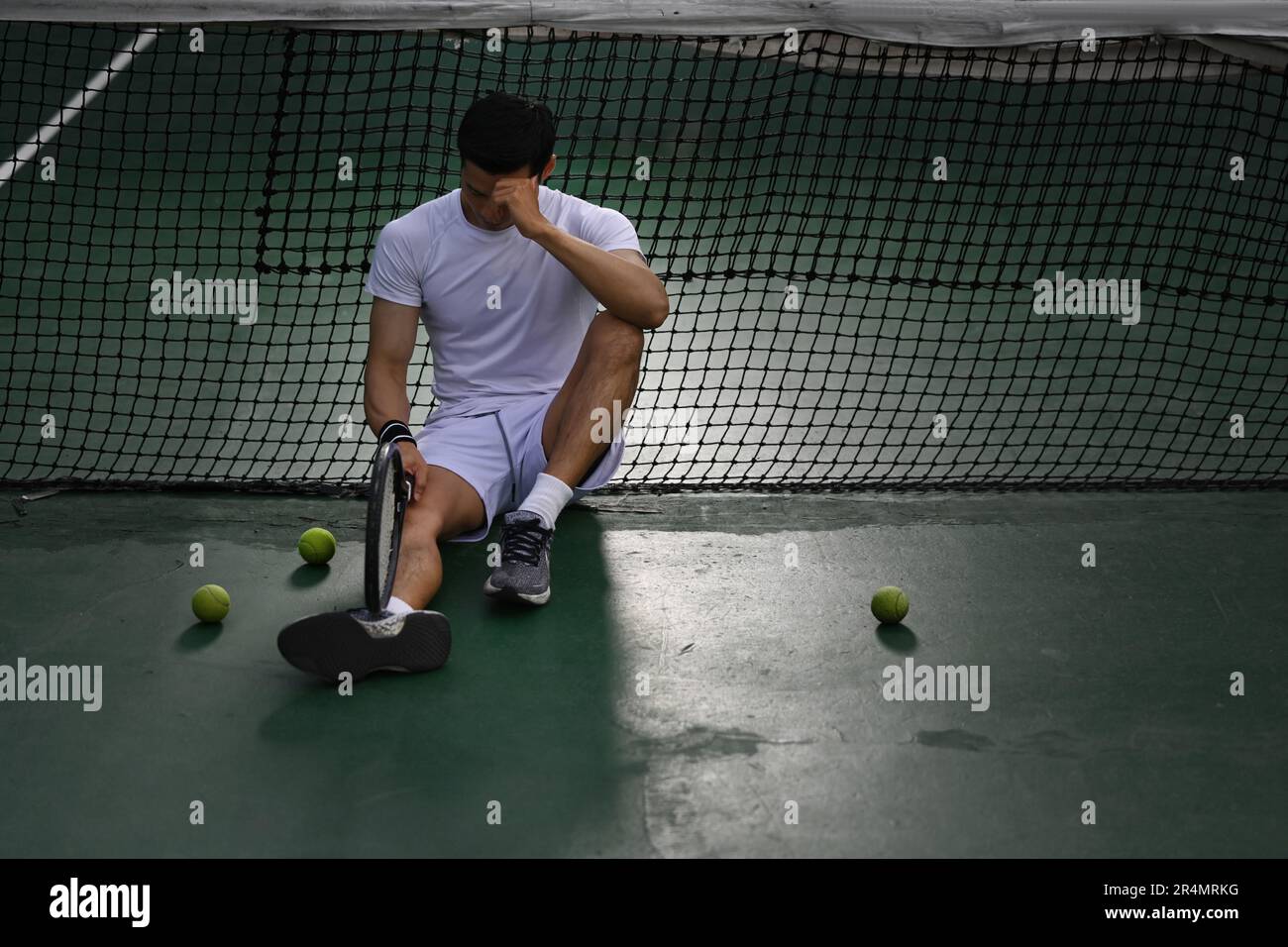 Giocatore asiatico di tennis maschile deluso seduto sul campo, turbato per la perdita di concorrenza, emozioni di fallimento Foto Stock