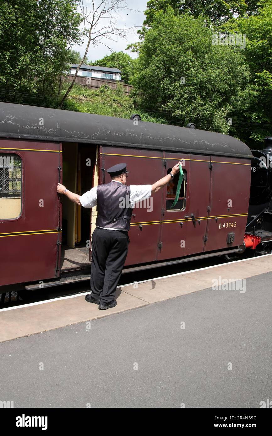 Guardia sulla Keighley & Worth Valley Heritage Railway segnala immediatamente con una bandiera verde prima di salire a bordo del treno Foto Stock