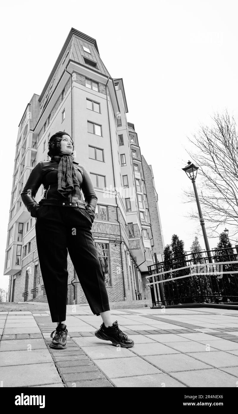 Una giovane e graziosa donna europea in un cappotto di pelle alla moda con pantaloni neri si trova vicino a un edificio vintage della città. Bella ragazza alla moda Foto Stock