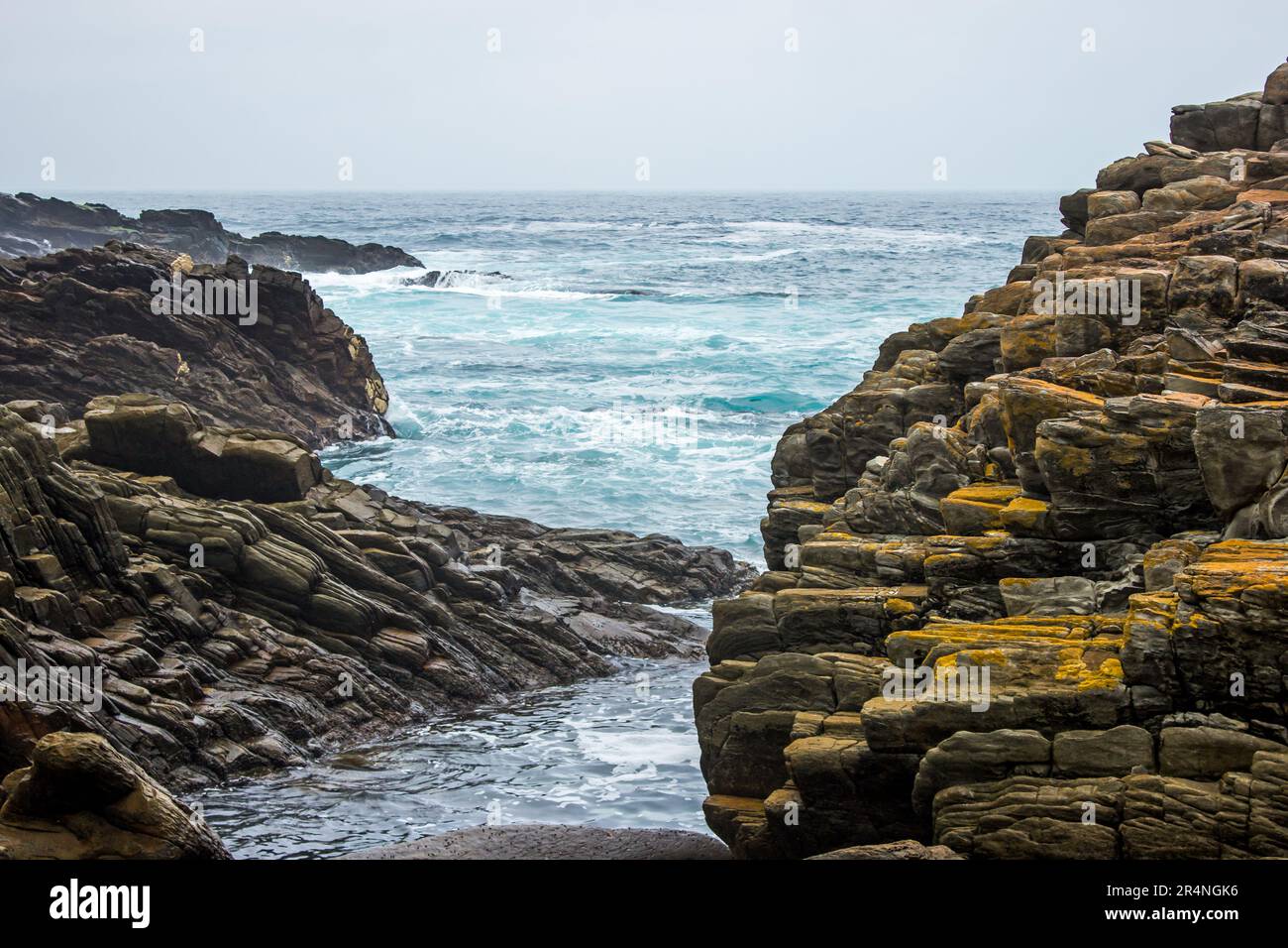 Guardando attraverso le rocce del tiltel della costa di Tsitsikama, Sud Africa, al grigio e ciano oceano di colore su una fredda mattina coperto Foto Stock