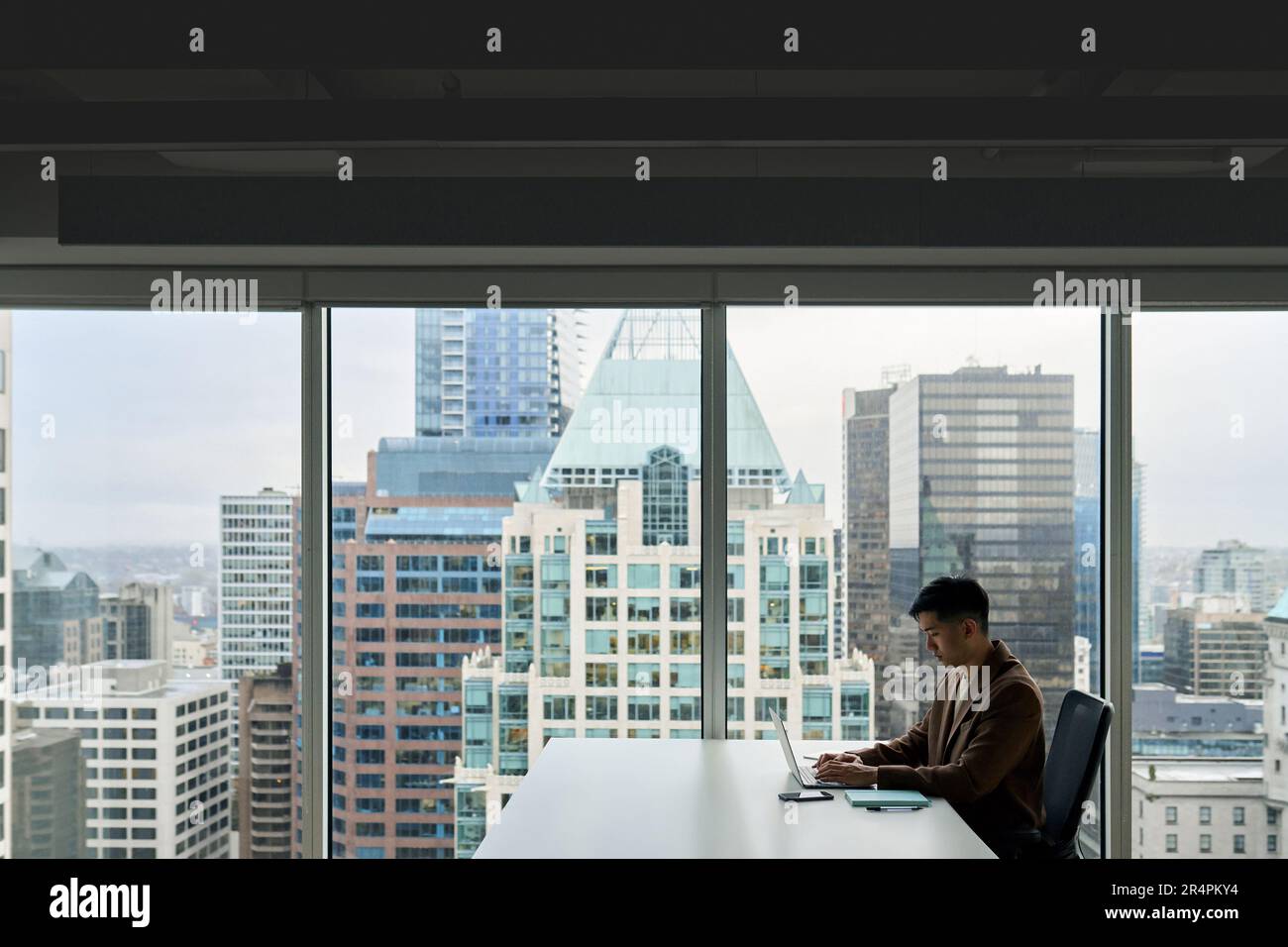Giovane uomo d'affari asiatico che lavora su un computer portatile in ufficio con una grande finestra. Foto Stock