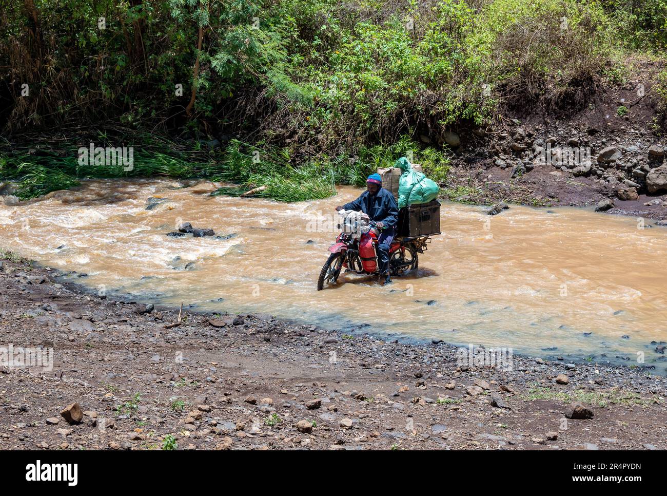 Un uomo locale su una motocicletta pesantemente caricata che cerca di attraversare un fiume. Kenya, Africa. Foto Stock