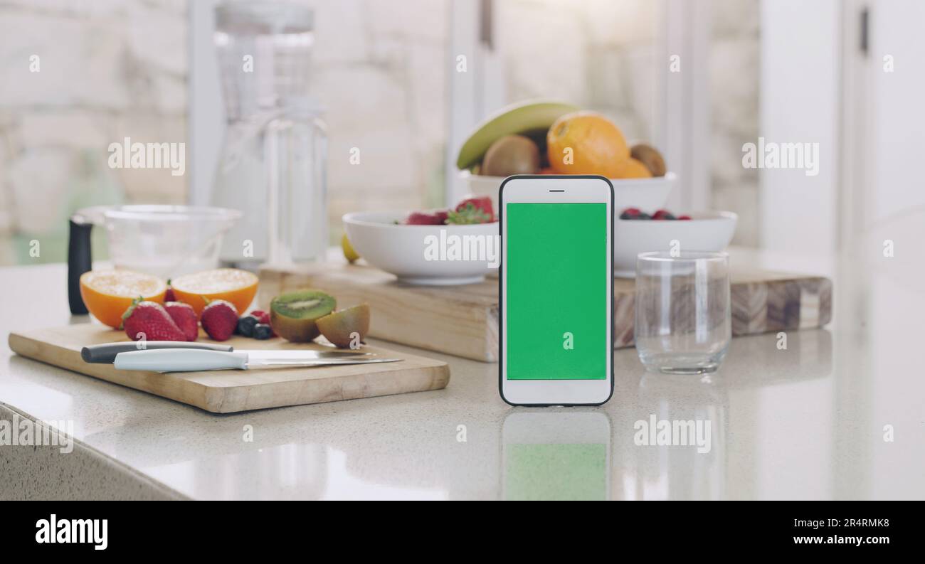 Telefono, schermo verde e cucina con frutta, dieta app e tavolo da sfondo sfocato in casa per frullato. Smartphone, bancone e frutta con spazio Foto Stock