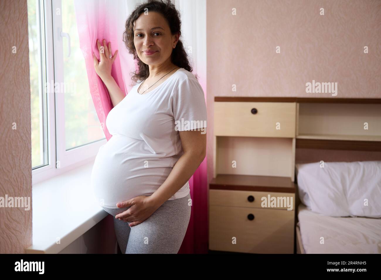 Donna incinta felice adulto in piedi alla finestra nella camera da letto domestica, toccante il suo ventre in gravidanza tardiva, sorridente alla macchina fotografica Foto Stock