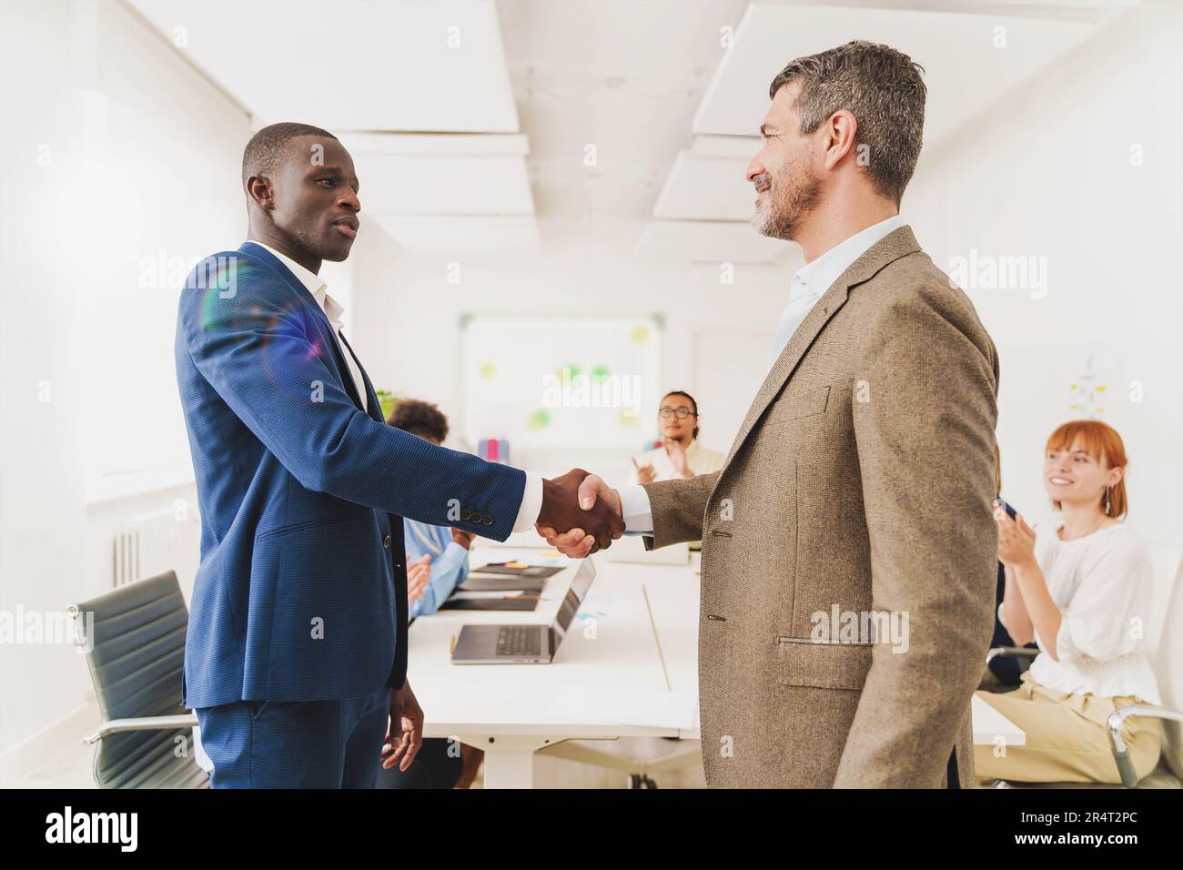 Multicultural Office Handshake - un africano e un caucasico che scuotono le mani in un ufficio moderno, mentre i colleghi applaudono in background. Foto Stock