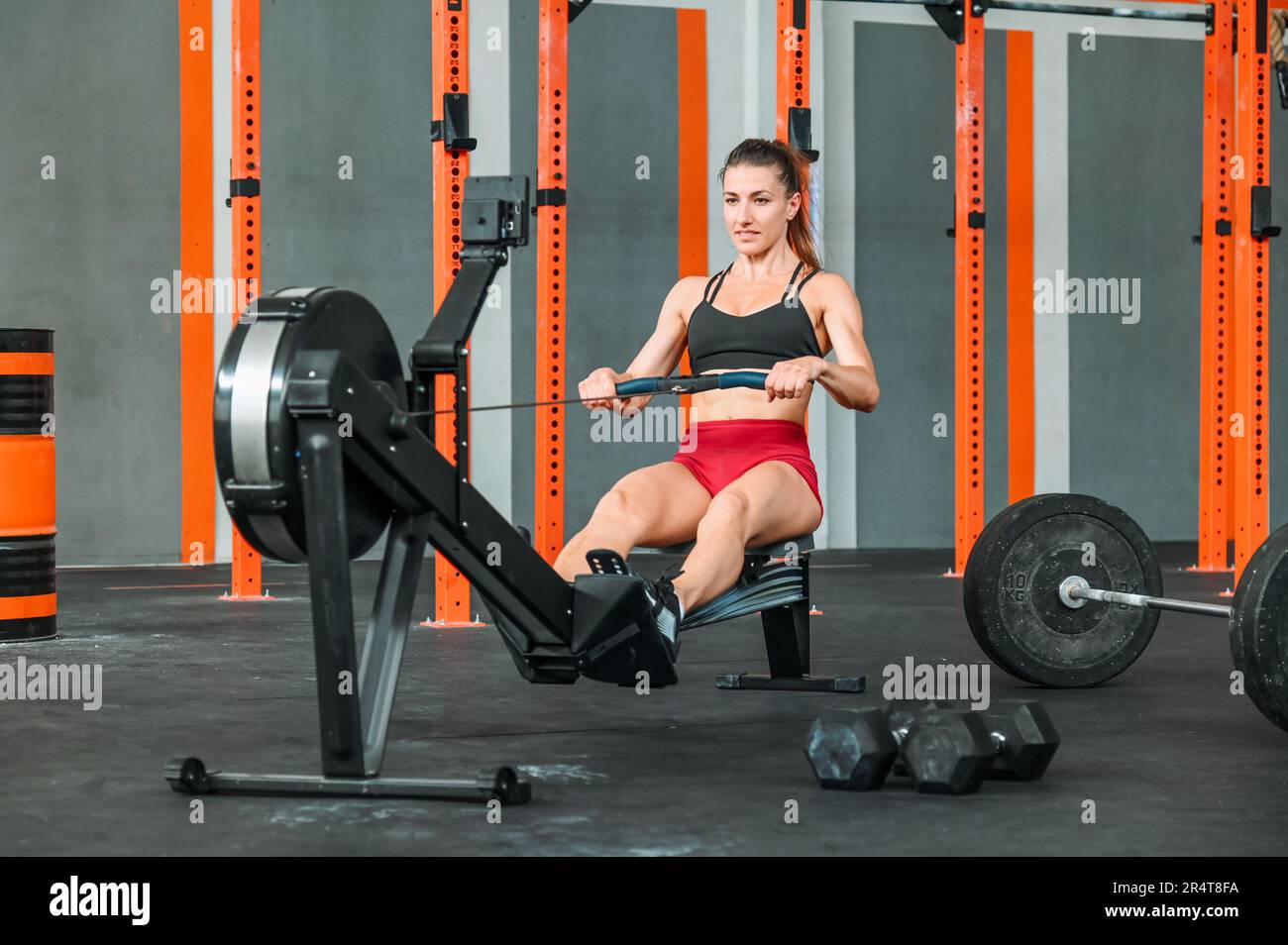 Corpo pieno di atleta giovane e forte con lunghi capelli scuri in abbigliamento sportivo che tira la corda e sorride mentre si esercita sulla macchina per cavi in moderno sp Foto Stock